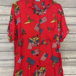 Puede incluir: Camisa roja con botones con un estampado patriótico con banderas americanas, la Estatua de la Libertad y águilas. La camisa de manga corta tiene un bolsillo en el pecho y botones negros. La camisa está colgada en una percha azul.