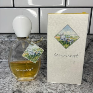 May include: A clear glass perfume bottle with a white rounded cap and golden liquid inside, featuring the word Summerset printed on the front and a diamond-shaped tag showing a blue chair. Beside it stands a tall white rectangular box with the same label.