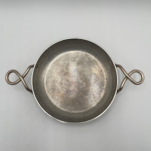 May include: A round metal pan with two decorative handles. The pan has a silver finish and a slightly textured surface. The handles are shaped in a figure-eight design. The pan is empty and sits on a white surface.