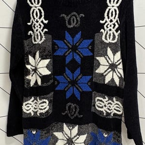Pode incluir: Um suéter preto de manga comprida com gola redonda e uma etiqueta com o nome Joyce. A peça apresenta um padrão de motivos geométricos de flocos de neve em azul, branco e cinza, além de intrincados desenhos bordados em branco.