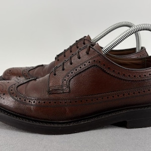 May include: A pair of dark brown leather wingtip oxford shoes featuring a textured pebble grain finish, decorative brogue detailing along the seams, thin dark laces, and a thick, solid black rubber sole with a low heel.