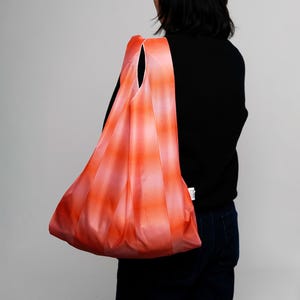 Op de afbeelding: Een herbruikbare boodschappentas met een roze en oranje streepjespatroon. De tas wordt over de schouder gedragen en is gemaakt van lichtgewicht materiaal. Een levendig accessoire voor het dragen van boodschappen of andere items.