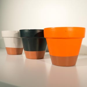 May include: Three small, painted terracotta pots. One is white, one is dark green, and one is bright orange. Each pot has a terracotta-colored base. The pots are arranged in a row against a white background.