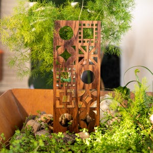 May include: A wooden decorative panel with geometric cutouts, including circles, squares, and diamond shapes. The panel is a warm brown color and is placed among greenery and small rocks, creating a natural aesthetic. The panel is approximately 12 inches tall.