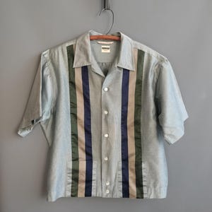 May include: Light blue short-sleeve shirt with vertical stripes in olive green, navy blue, and tan. The shirt has a button-down front, two front pockets, and a collar. The shirt is hanging on a wooden hanger.