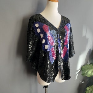 May include: A black sequined top featuring a large butterfly design in shades of blue, pink, and white. The butterfly has a beaded centre.