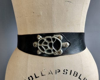 May include: A black leather belt with a silver turtle buckle. The buckle is detailed with a cutout design and has a textured finish.