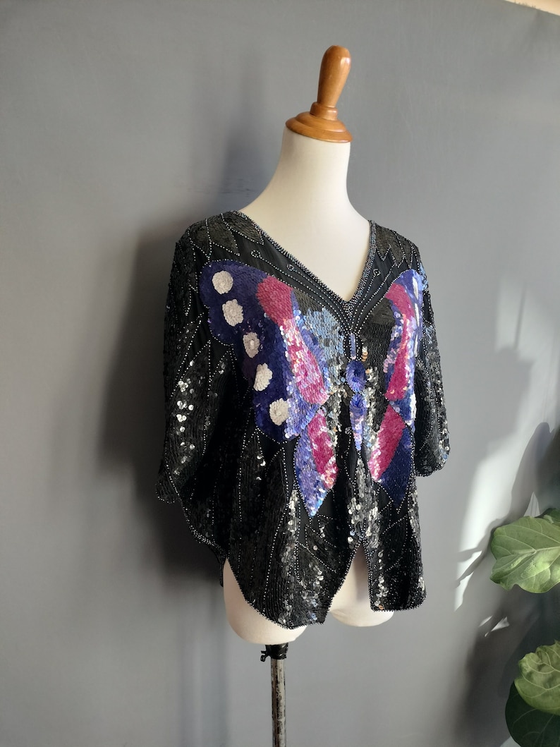 May include: A black sequined top featuring a large butterfly design in shades of blue, pink, and white. The butterfly has a beaded centre.