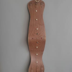 May include: A brown leather horse saddle pad with decorative embossed floral design and metal studs. The pad is suspended from a gold-colored hook. The pad has leather straps and buckles.
