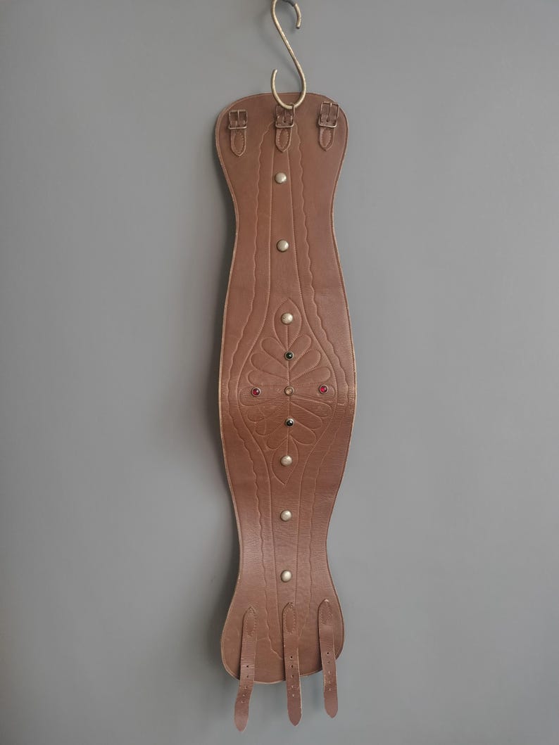 May include: A brown leather horse saddle pad with decorative embossed floral design and metal studs. The pad is suspended from a gold-colored hook. The pad has leather straps and buckles.