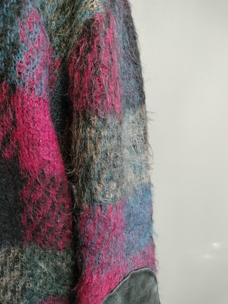 May include: Close-up of a textured, multi-colored sweater. The sweater features a blend of pink, blue, teal, and gray hues. The fabric appears soft and fuzzy, with a patch of gray material at the bottom. The overall style is vintage.
