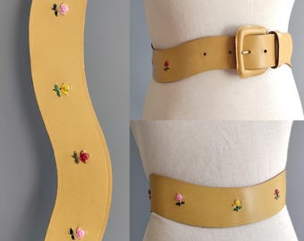 May include: Tan leather belt with a large square buckle. The belt features embroidered floral accents in pink, yellow, and red. The belt is displayed on a mannequin.