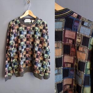 May include: A long-sleeve crew neck sweater featuring an all-over jacquard knit pattern of multi-colored floppy disks in shades of blue, green, black, and brown. The garment has a ribbed collar, cuffs, and hem with thin, dark, multi-colored stripes.