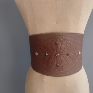 May include: A wide brown leather belt with a floral embossed design and decorative silver studs. The belt features red and green jewel accents and is displayed on a mannequin.