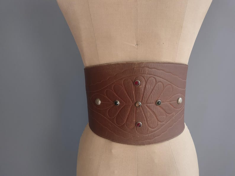 May include: A wide brown leather belt with a floral embossed design and decorative silver studs. The belt features red and green jewel accents and is displayed on a mannequin.