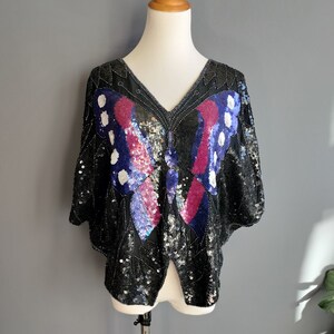 May include: A black sequined top featuring a butterfly design in shades of pink, blue, and white. The butterfly is positioned centrally on the front of the top.
