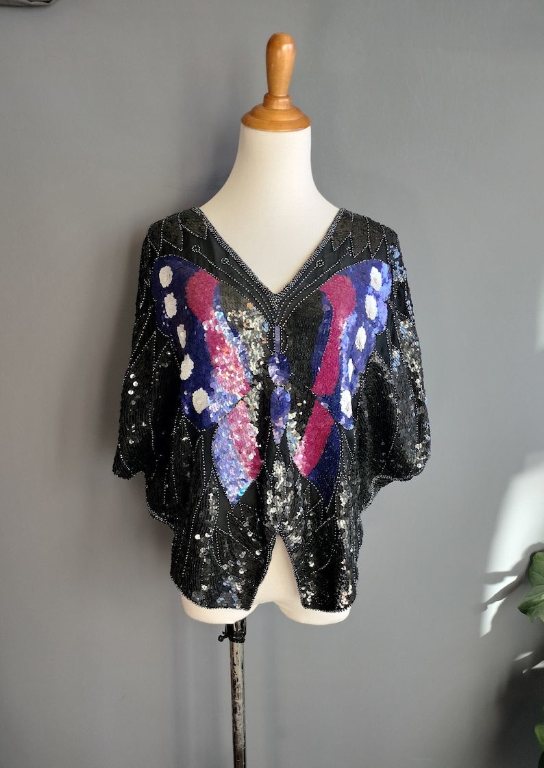 May include: A black sequined top featuring a butterfly design in shades of pink, blue, and white. The butterfly is positioned centrally on the front of the top.