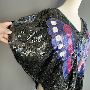 May include: A black sequined top featuring a butterfly design in shades of purple, pink, and white. The top has a V-neck and short sleeves.