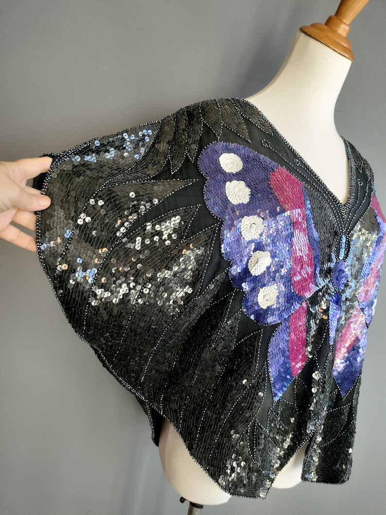 May include: A black sequined top featuring a butterfly design in shades of purple, pink, and white. The top has a V-neck and short sleeves.
