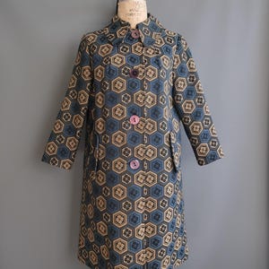 May include: A vintage knee-length coat with a geometric pattern in shades of blue, tan, and black. The coat features a collared neckline, long sleeves, and large, round buttons in shades of purple and pink. The coat is displayed on a mannequin.