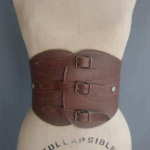 May include: A brown leather corset belt with three buckle closures and decorative stitching. The wide belt is displayed on a mannequin, showcasing its shape and design. The belt has a vintage aesthetic.