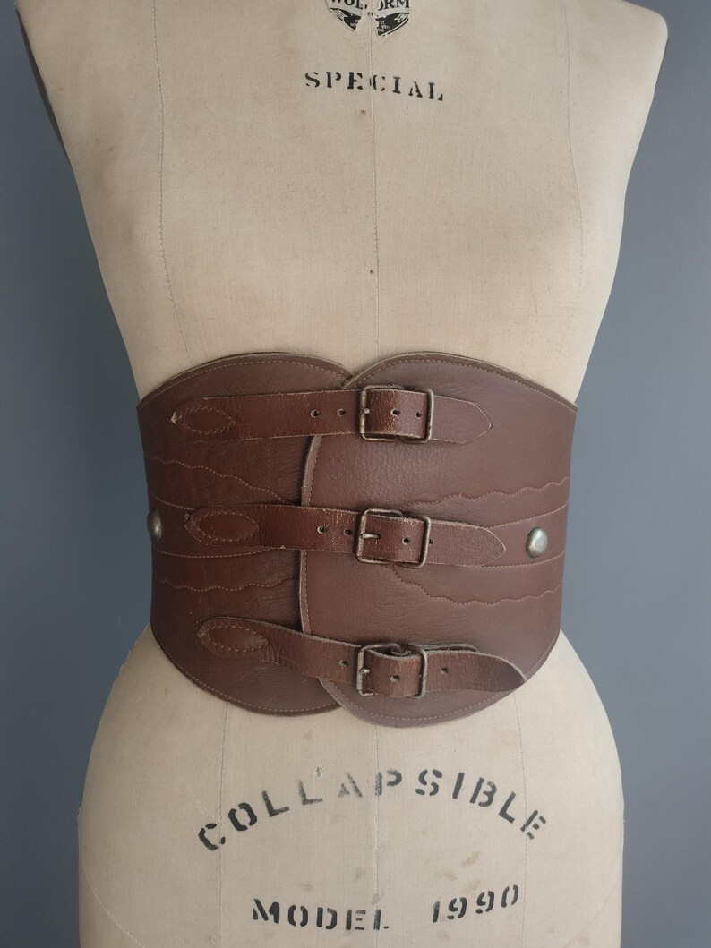 May include: A brown leather corset belt with three buckle closures and decorative stitching. The wide belt is displayed on a mannequin, showcasing its shape and design. The belt has a vintage aesthetic.