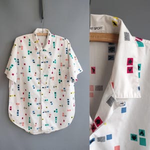 May include: A white short-sleeve button-down shirt featuring a repeating pattern of small colorful squares, rectangles, and graphic icons including a rooster and house. The collar tag displays the text "SPIRIT SPORT" in black capital letters.