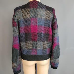 May include: A vintage, oversized sweater in a plaid pattern of gray, black, blue, and magenta. The sweater has black suede elbow patches and a black ribbed hem. The garment is displayed on a mannequin.