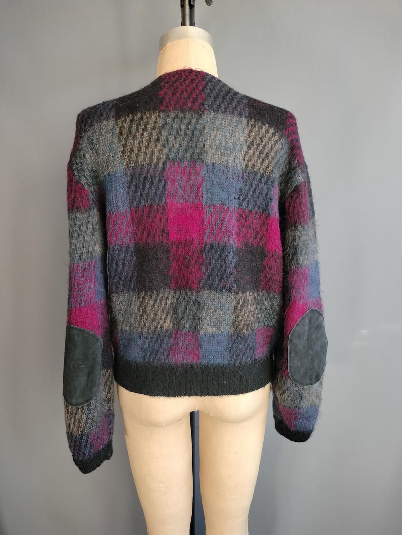 May include: A vintage, oversized sweater in a plaid pattern of gray, black, blue, and magenta. The sweater has black suede elbow patches and a black ribbed hem. The garment is displayed on a mannequin.