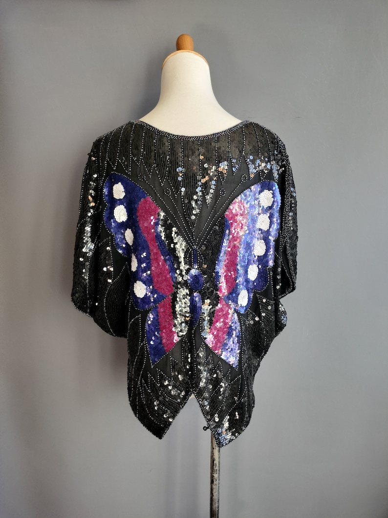 May include: A black sequined top featuring a large butterfly design in shades of blue, pink, and white.