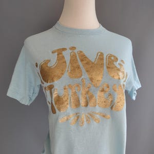 May include: Pale blue t-shirt featuring the words "Jive Turkey" in gold lettering. The text is in a retro, rounded font. The short-sleeved shirt is made of a soft material. The t-shirt is displayed on a mannequin.