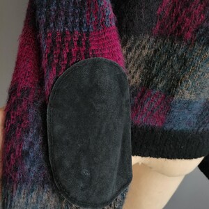 May include: A close-up of a sweater sleeve with a black suede elbow patch. The sweater features a plaid pattern in shades of pink, blue, black, and brown. The cuff is black.