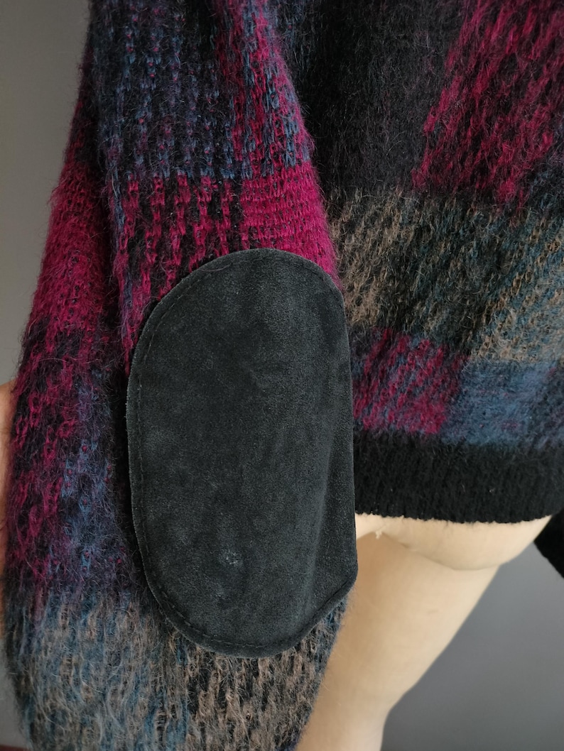 May include: A close-up of a sweater sleeve with a black suede elbow patch. The sweater features a plaid pattern in shades of pink, blue, black, and brown. The cuff is black.