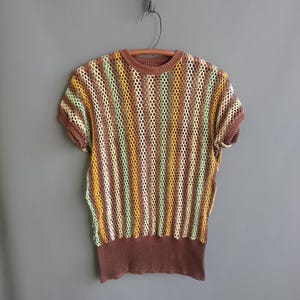 May include: A short-sleeved, open-weave top in a brown, yellow, and green striped pattern. The top has a brown ribbed neckline, short sleeves, and a ribbed hem. The garment is hanging on a wooden hanger against a gray background.