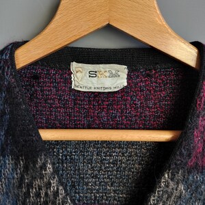May include: A multi-colored knitted cardigan with a button closure. The garment features a mix of blue, black, and burgundy hues. The label reads "SKM SEATTLE KNITTING MILLS". The cardigan is displayed on a wooden hanger.