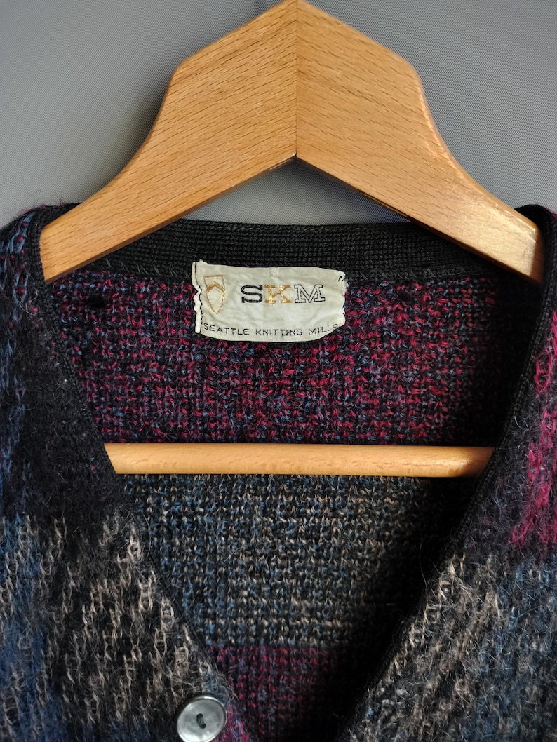 May include: A multi-colored knitted cardigan with a button closure. The garment features a mix of blue, black, and burgundy hues. The label reads "SKM SEATTLE KNITTING MILLS". The cardigan is displayed on a wooden hanger.