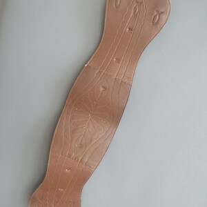 May include: A brown leather corset-style belt with decorative floral and linear embossed patterns. The belt has a long, curved shape with a hook at the top and three small tabs at the bottom. The belt is shown against a gray background.