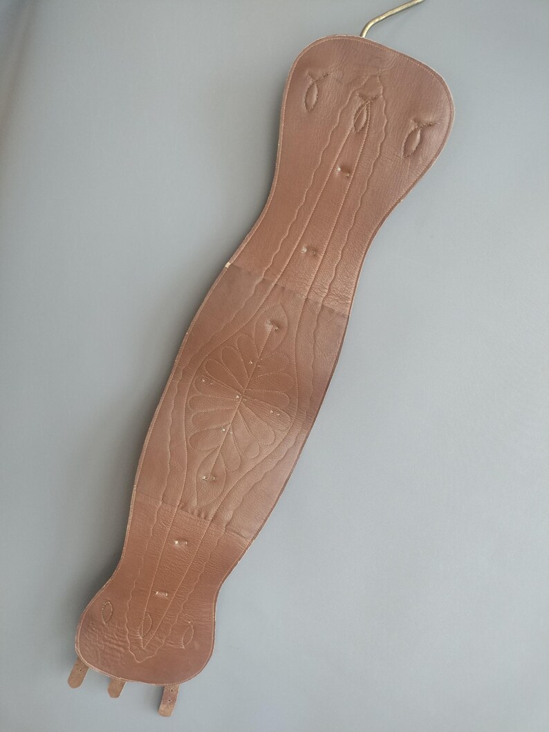May include: A brown leather corset-style belt with decorative floral and linear embossed patterns. The belt has a long, curved shape with a hook at the top and three small tabs at the bottom. The belt is shown against a gray background.