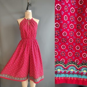 Könnte beinhalten: Ein roter Vintage-Halter-Jumpsuit mit floralem Muster. Der Jumpsuit hat einen Schlüsselloch-Ausschnitt, eine elastische Taille und weite Beine. Der Stoff hat ein sich wiederholendes Muster aus kleinen weißen, blauen und schwarzen Blumen. Der Saum ist mit einer dekorativen Bordüre versehen.