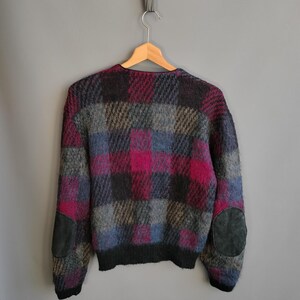 May include: A vintage sweater with a plaid pattern in shades of pink, gray, blue, and black. The sweater has dark gray suede elbow patches and a black ribbed hem and cuffs. The sweater is hanging on a wooden hanger.