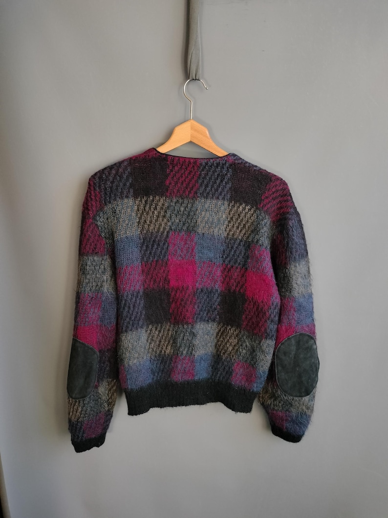 May include: A vintage sweater with a plaid pattern in shades of pink, gray, blue, and black. The sweater has dark gray suede elbow patches and a black ribbed hem and cuffs. The sweater is hanging on a wooden hanger.