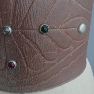May include: Close-up of a brown leather corset with decorative stitching and gemstone accents. The corset features a leaf-like pattern and is adorned with silver-toned studs and colorful gems.