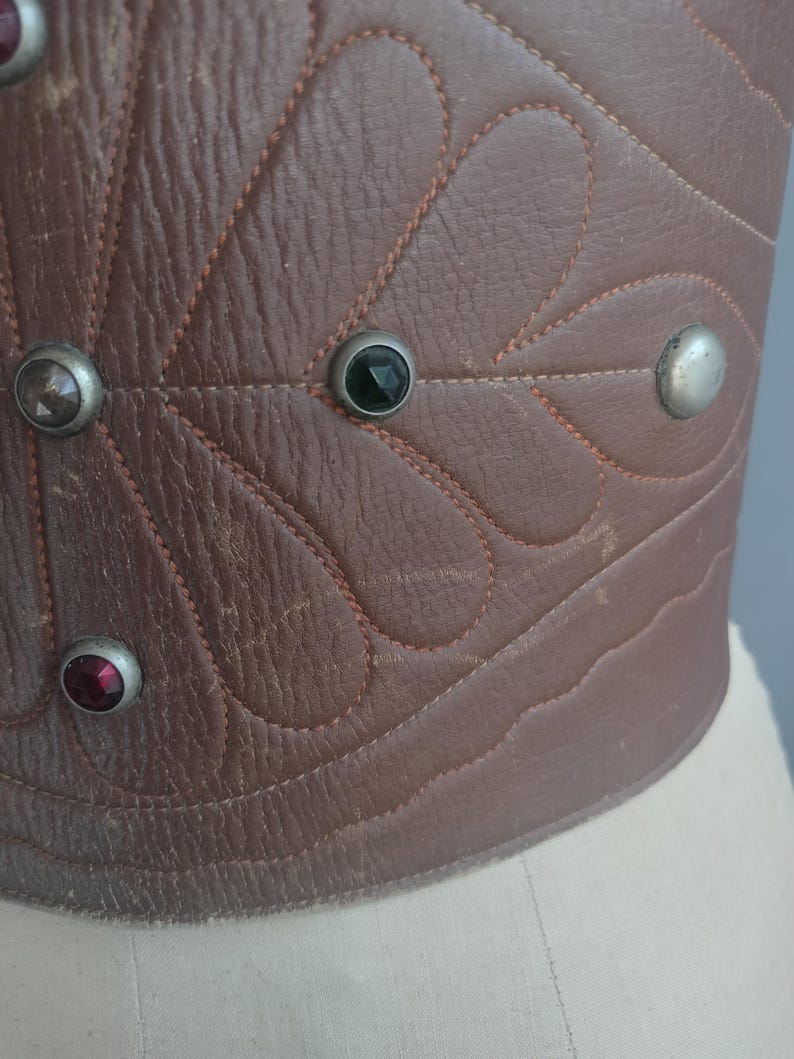 May include: Close-up of a brown leather corset with decorative stitching and gemstone accents. The corset features a leaf-like pattern and is adorned with silver-toned studs and colorful gems.
