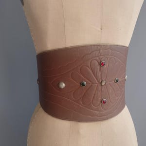 May include: A wide brown leather belt with an embossed floral design and decorative silver studs. The belt features red and green jewel accents. The belt is displayed on a mannequin.
