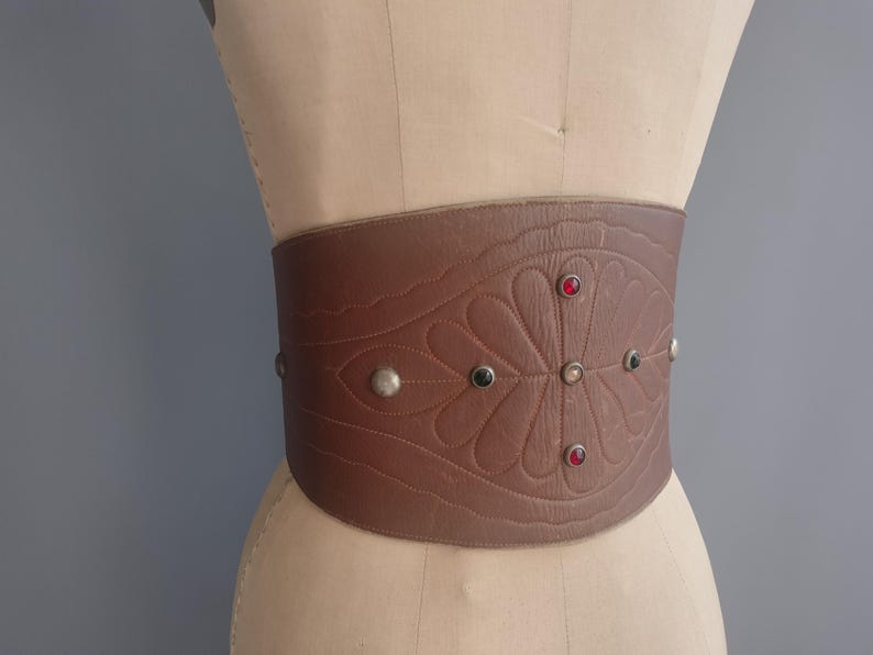May include: A wide brown leather belt with an embossed floral design and decorative silver studs. The belt features red and green jewel accents. The belt is displayed on a mannequin.