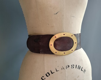 May include: A wide dark brown belt featuring a large oval-shaped brass buckle. The buckle is accented with small, evenly spaced red circular inlays around its perimeter. The belt strap is smooth and wraps around the waist of a dress form.