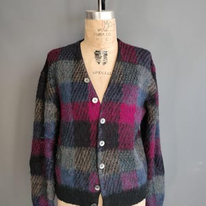 May include: A vintage, button-down cardigan sweater with a plaid pattern in shades of black, gray, blue, and magenta. The sweater is displayed on a mannequin and features a V-neckline and long sleeves. The buttons are white.