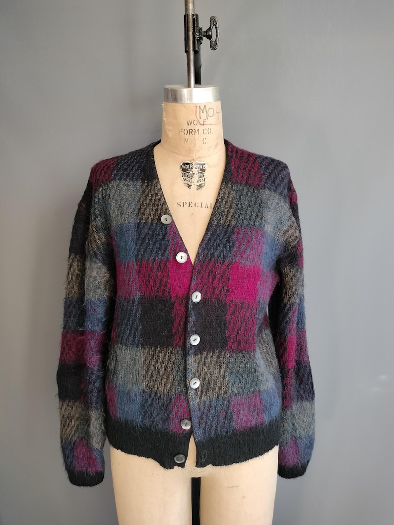 May include: A vintage, button-down cardigan sweater with a plaid pattern in shades of black, gray, blue, and magenta. The sweater is displayed on a mannequin and features a V-neckline and long sleeves. The buttons are white.