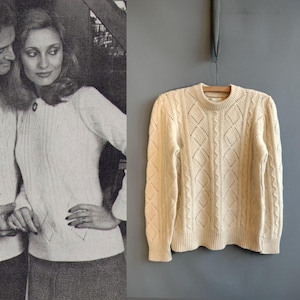 Puede incluir: Suéter de punto trenzado color crema con calados en forma de diamante, colgado de una percha de madera contra una pared gris. El suéter tiene cuello redondo y puños y dobladillo acanalados. A la izquierda hay una foto en blanco y negro de una pareja.