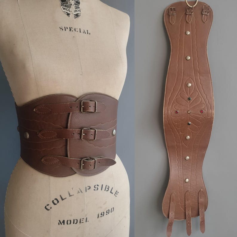 May include: Brown leather corset with three buckle closures and decorative stitching. The corset is displayed on a mannequin. A separate, matching leather piece with decorative rivets and a ring is also shown.